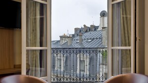 A room with two brown chairs, a small round table, and a view of a balcony with a wrought iron railing. - Hotel de Sevigne (Paris)
