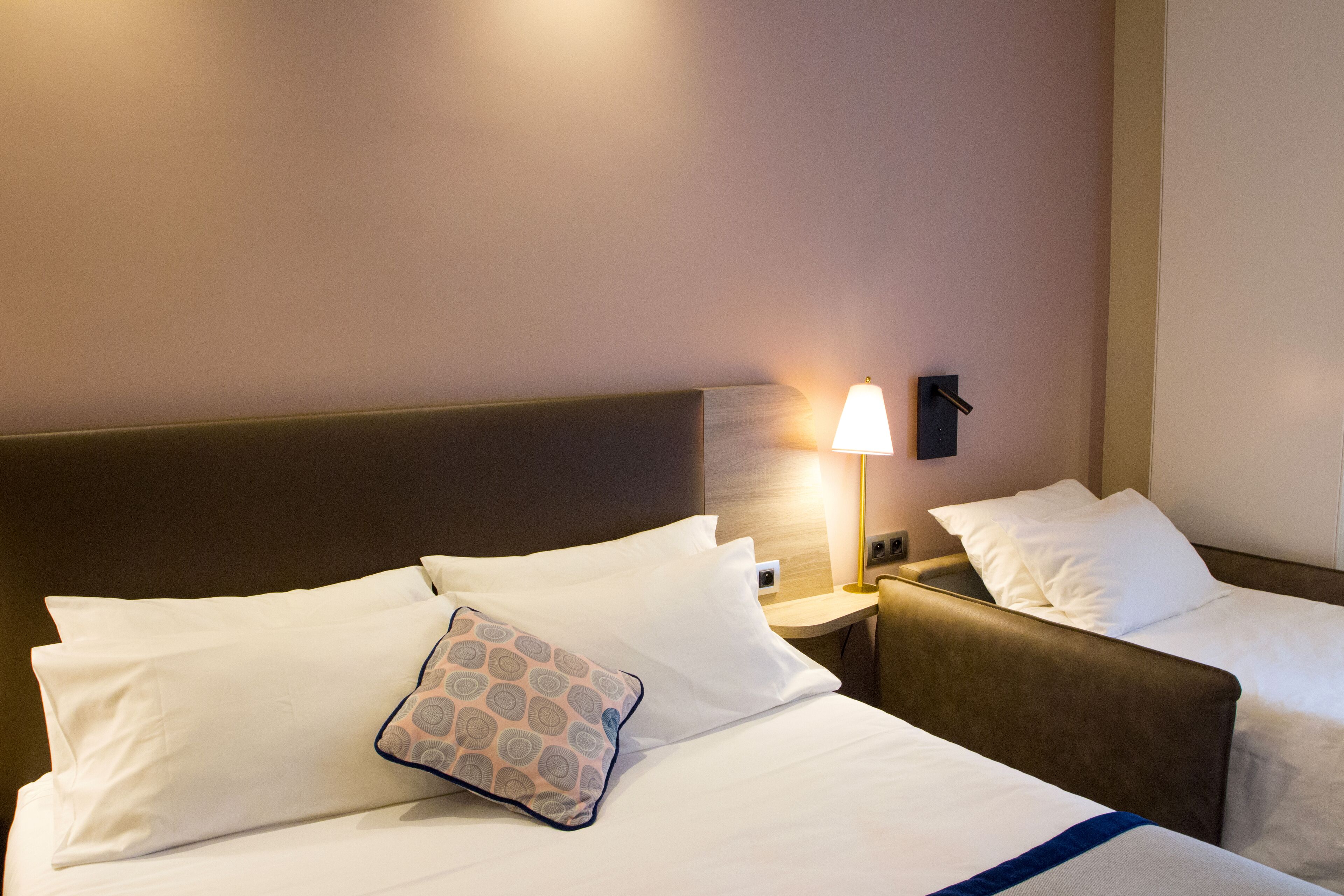 Et hotelværelse med to senge, et natbord med lampe og vægmonteret lys.