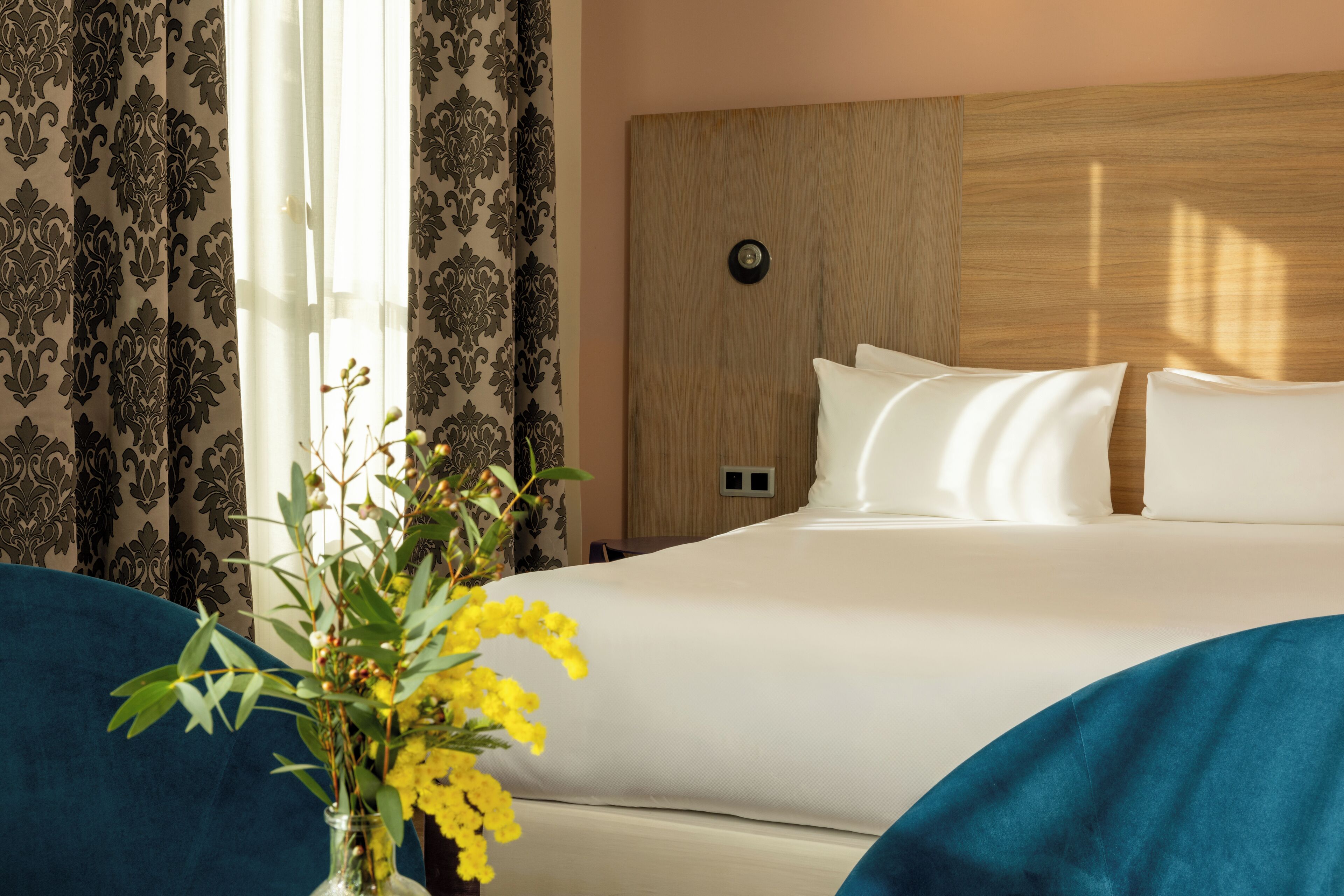 A hotel room with a large bed, a wooden headboard, and a vase with yellow flowers on a blue sofa.