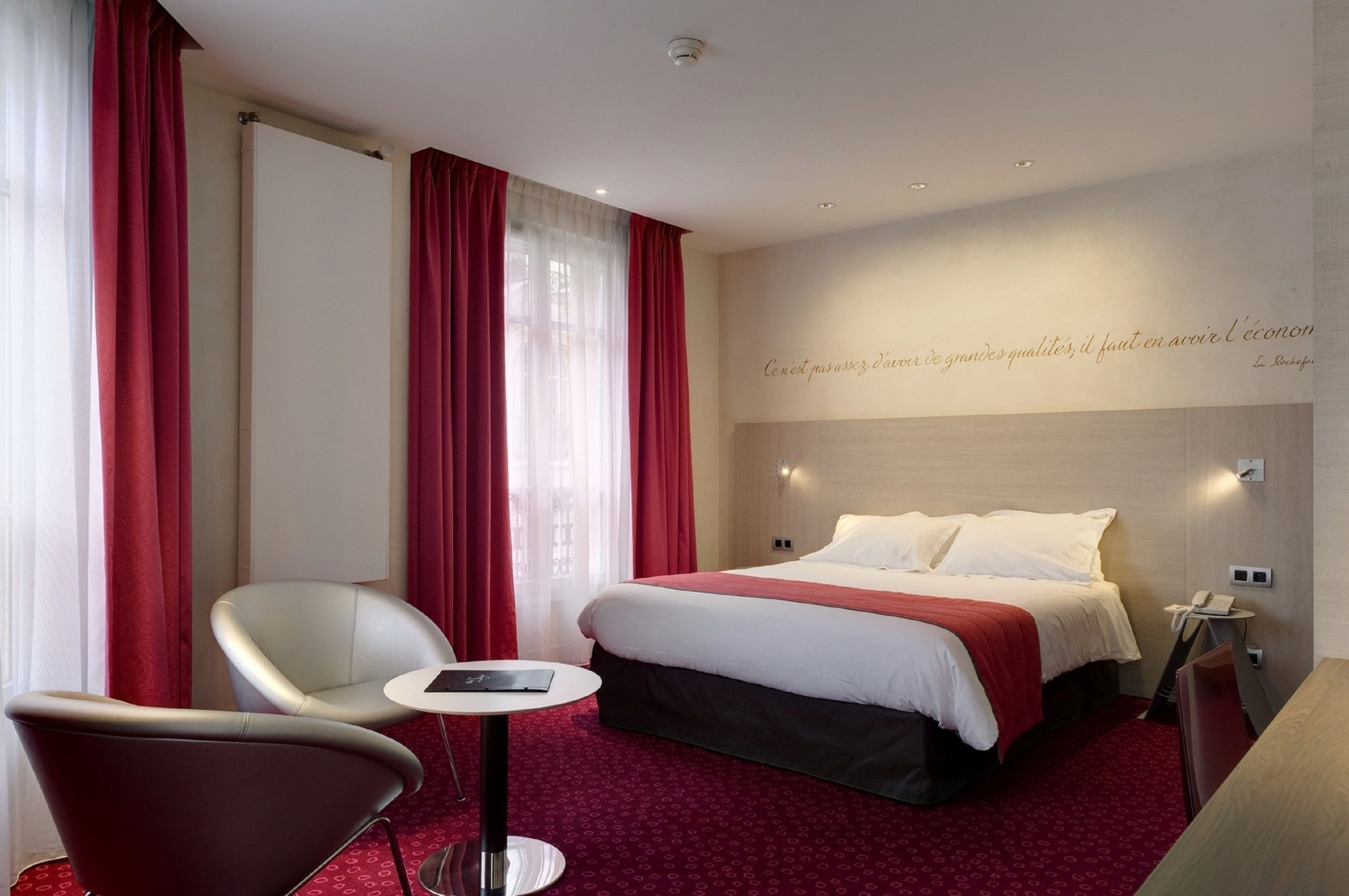 Une chambre d’hôtel avec un lit, une chaise, une petite table et un mur orné d’une citation.