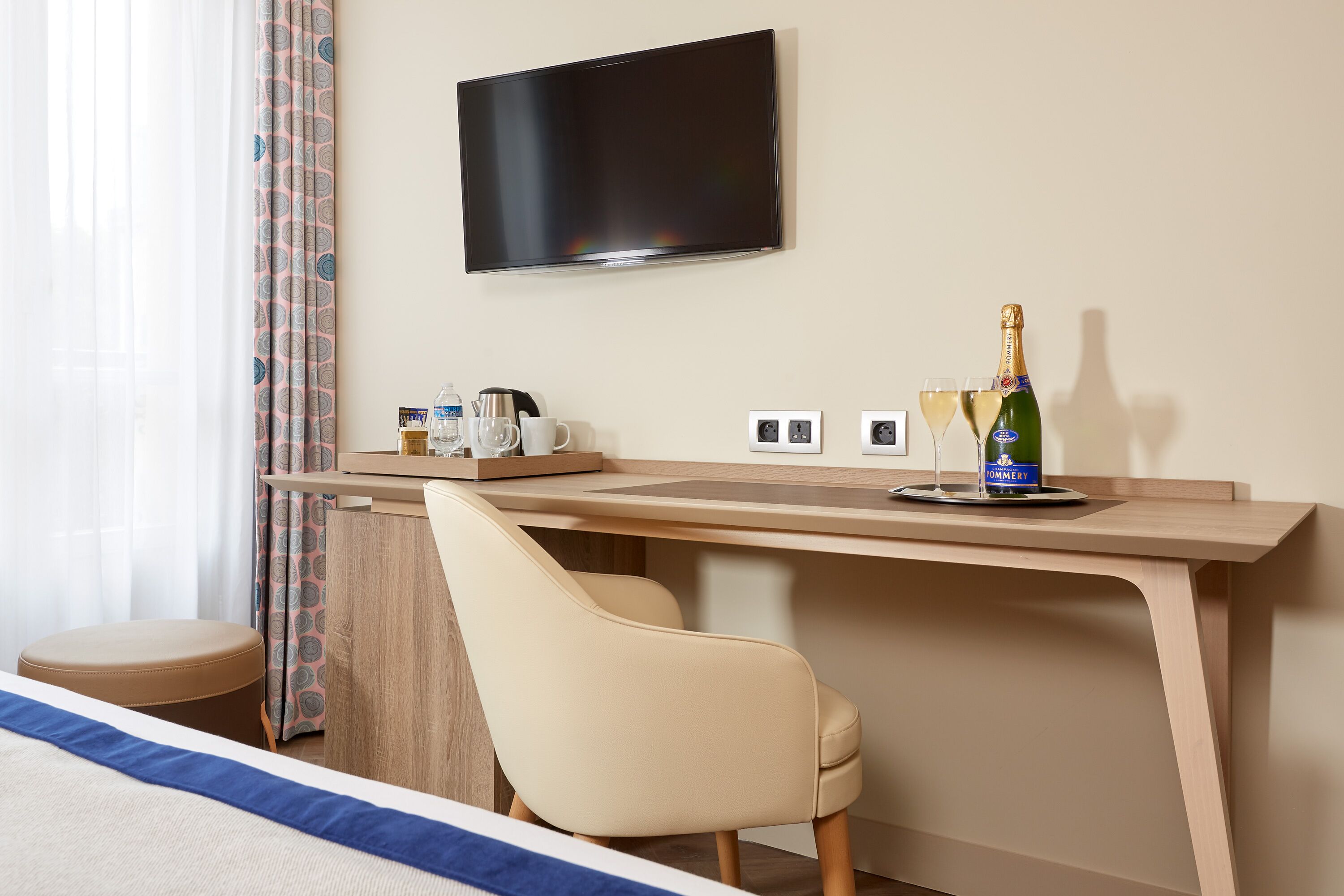 A hotel room with a bed, a desk with a chair, a television, a bottle of champagne, and glasses.