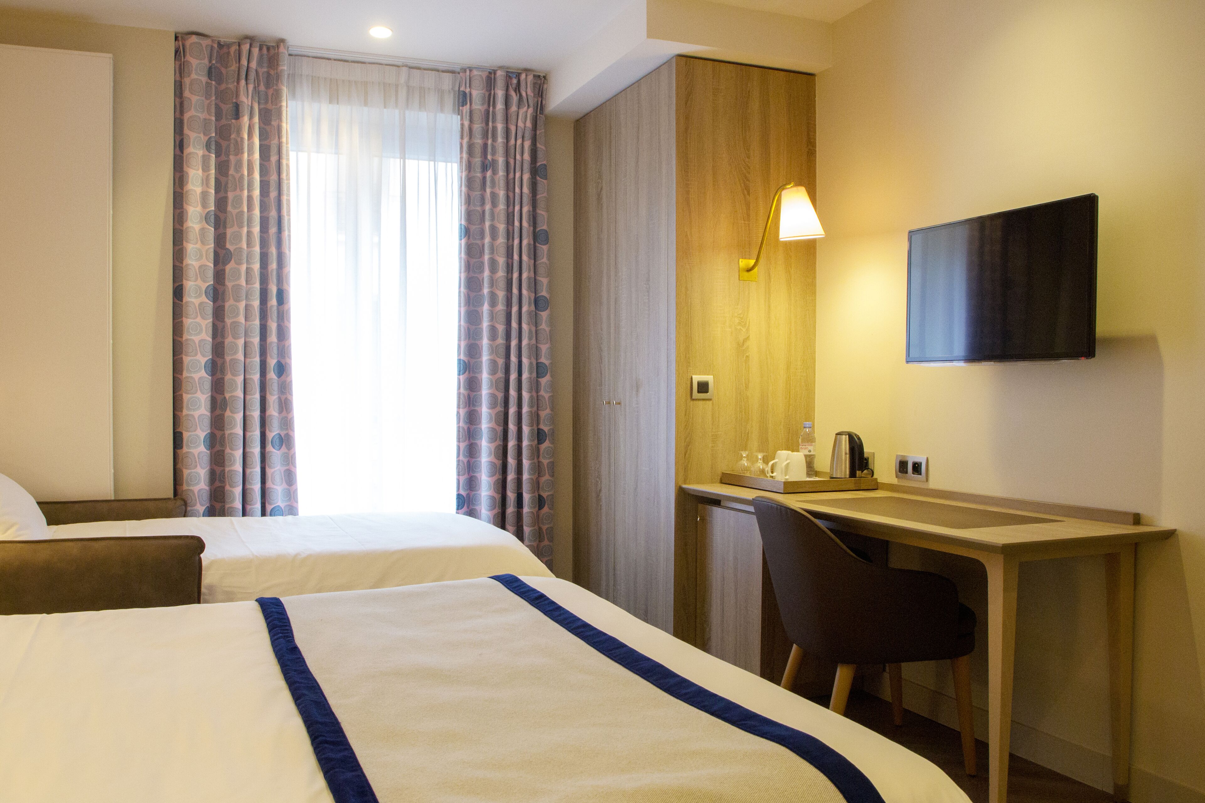 A hotel room with two beds, a desk with a chair, a flat-screen TV, and a window with curtains.