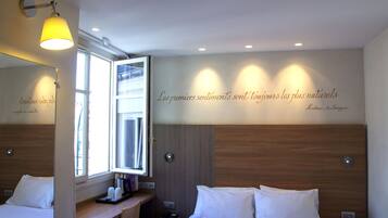 Een hotelkamer met een bed, nachtkastje, lamp en een muur met een Frans citaat.