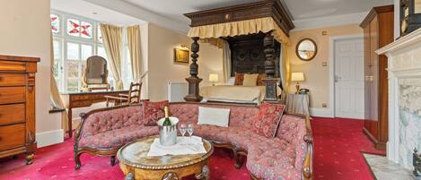 A traditional room with a four-poster bed, a red carpeted floor, a wooden coffee table with a pitcher and glasses, and a large mirror on the wall.