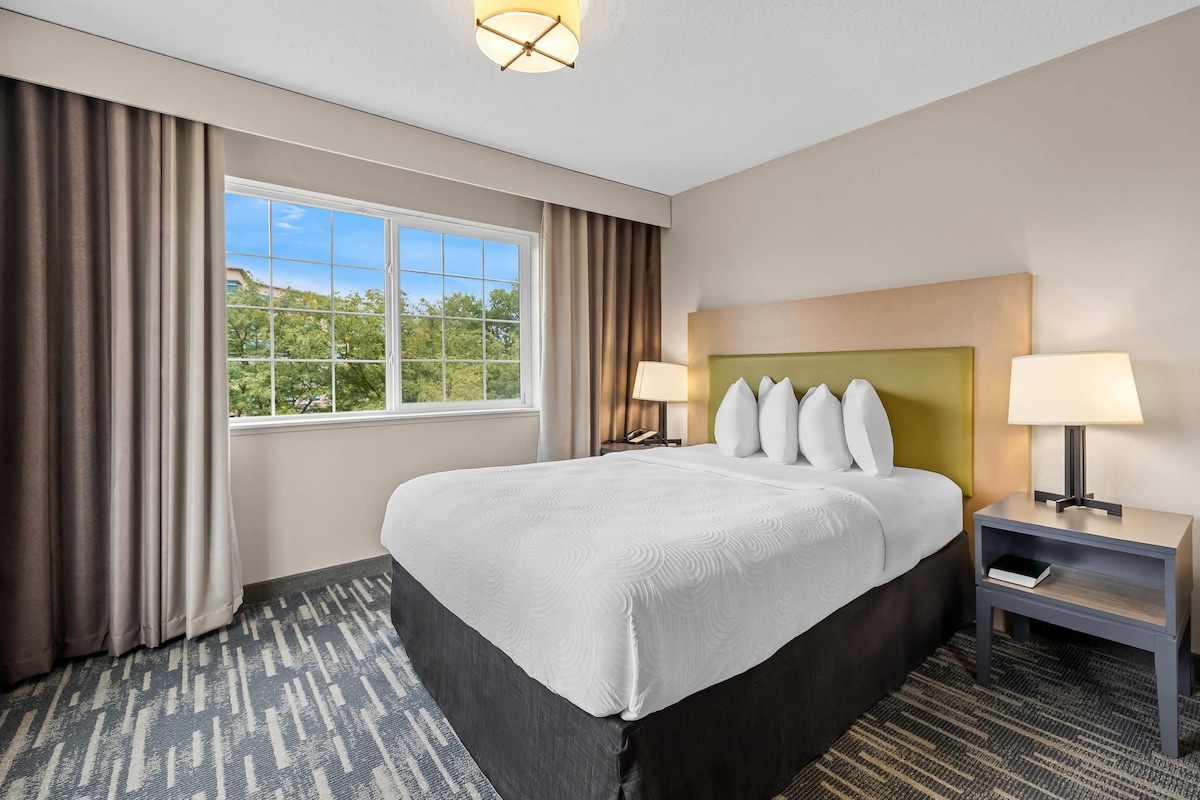 Quarto de hotel com uma cama grande, duas luminárias de cabeceira, uma mesa de cabeceira, uma janela com cortinas e vista para o verde lá fora.