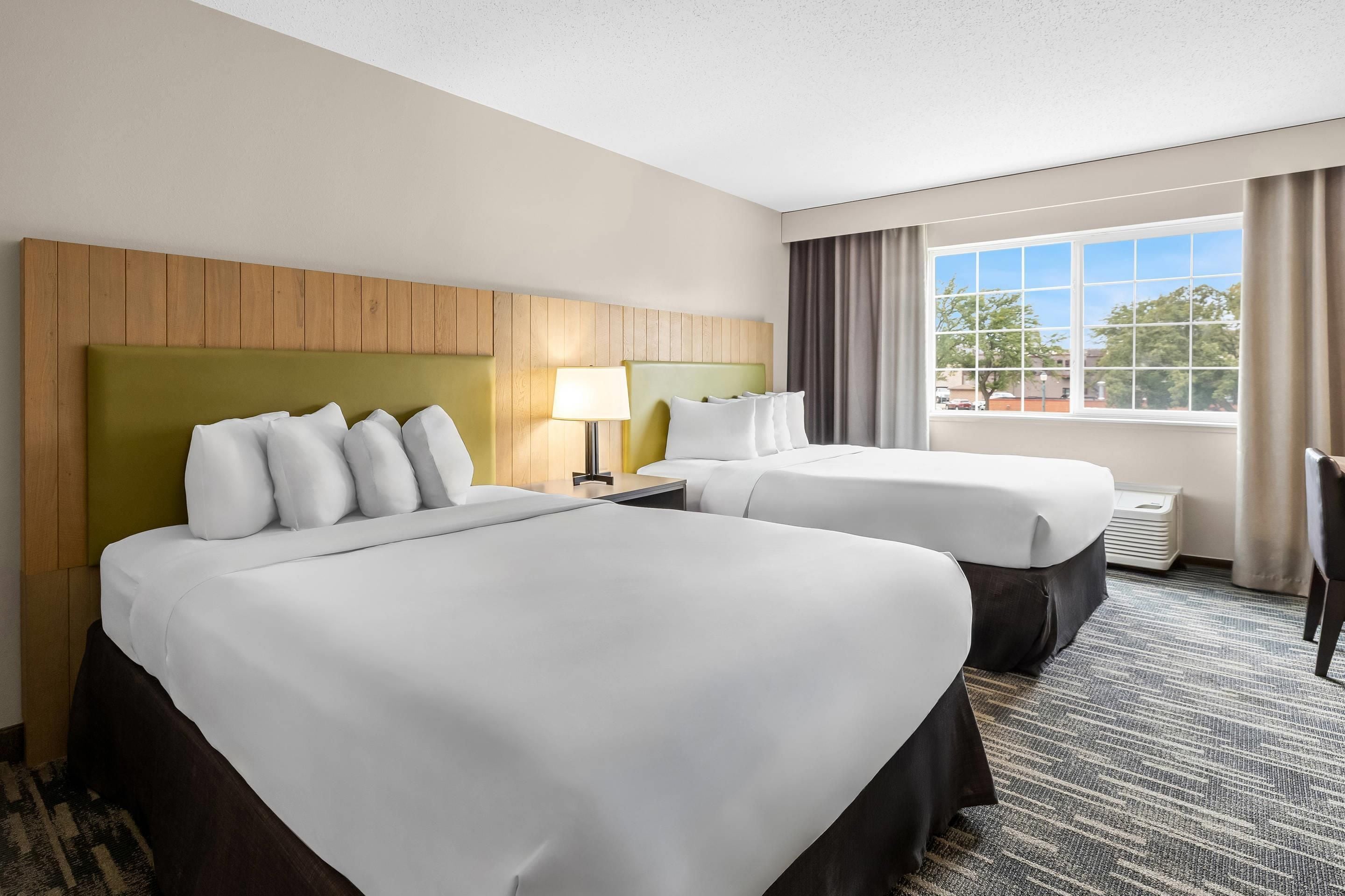 A hotel room with two beds, a nightstand, a lamp, and a view of the outdoors.