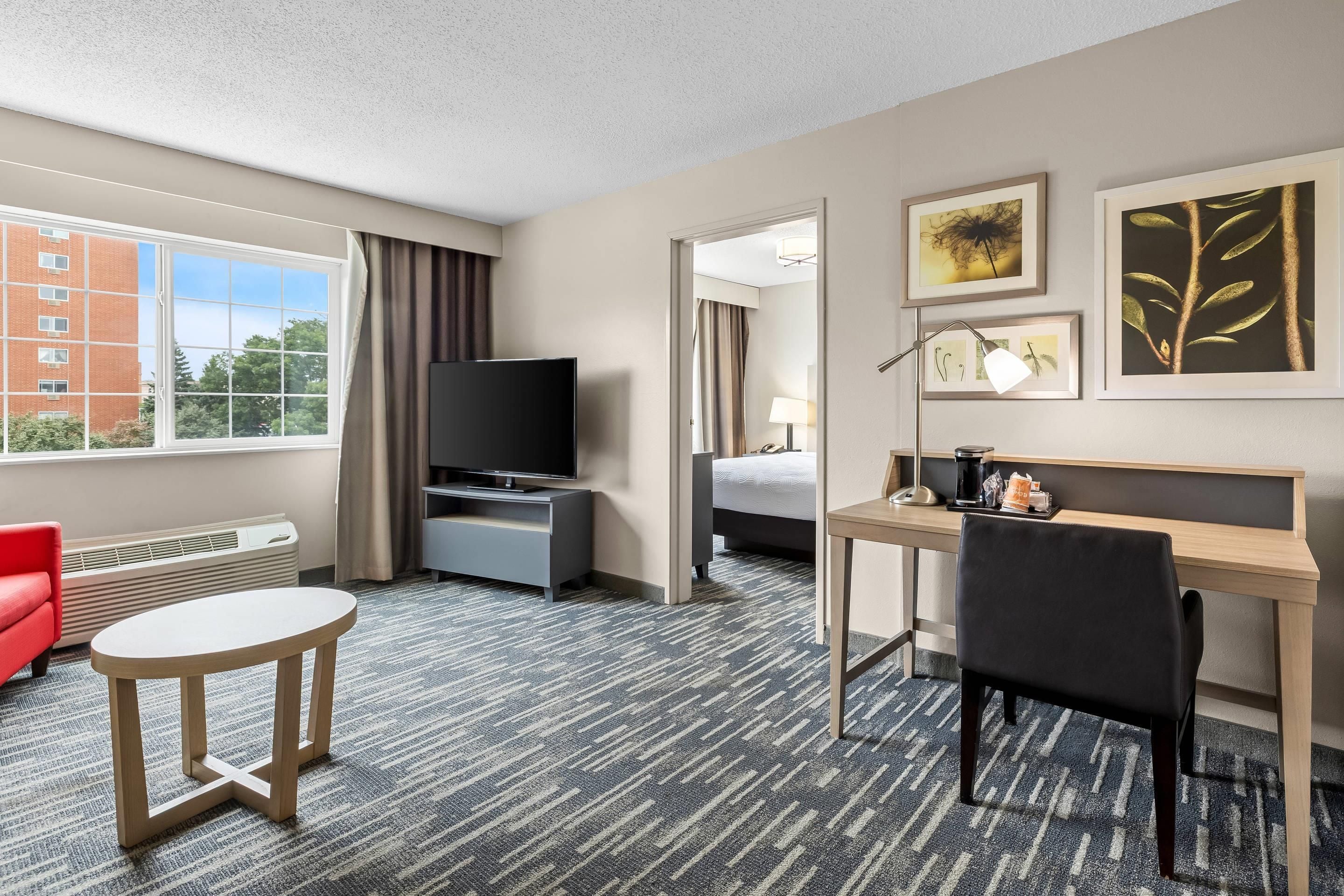 A hotel room with a bed, desk, chair, television, and a view of a building outside.