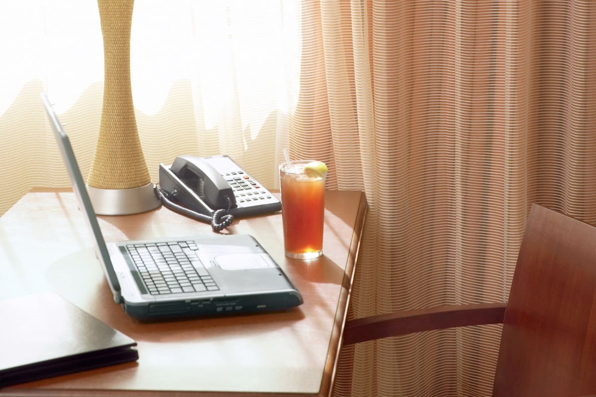 Quarto de hotel com uma mesa de madeira, um laptop, um telefone, um copo de suco de laranja, uma luminária e uma cadeira.