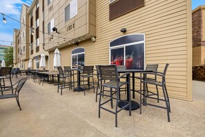 An outdoor seating area with tables and chairs, a building with beige siding, and a window displaying a drink.