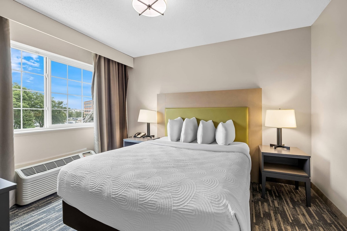 Quarto de hotel com uma cama grande, duas mesas de cabeceira com luminárias, uma janela com cortinas e vista para o exterior.
