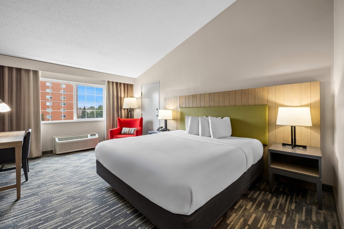 Quarto de hotel com uma cama grande, escrivaninha, cadeira e vista para um edifício pela janela.