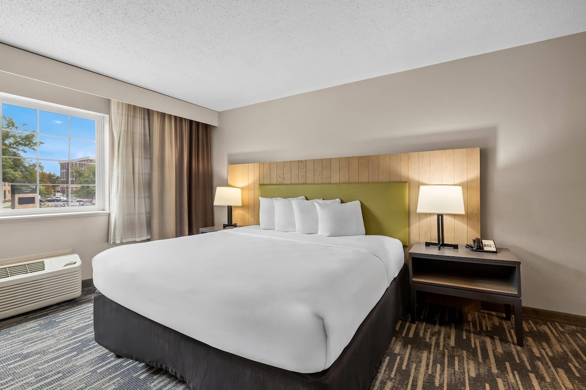 Quarto de hotel com uma cama grande, duas mesas de cabeceira com luminárias, uma janela com cortinas e vista para o exterior.
