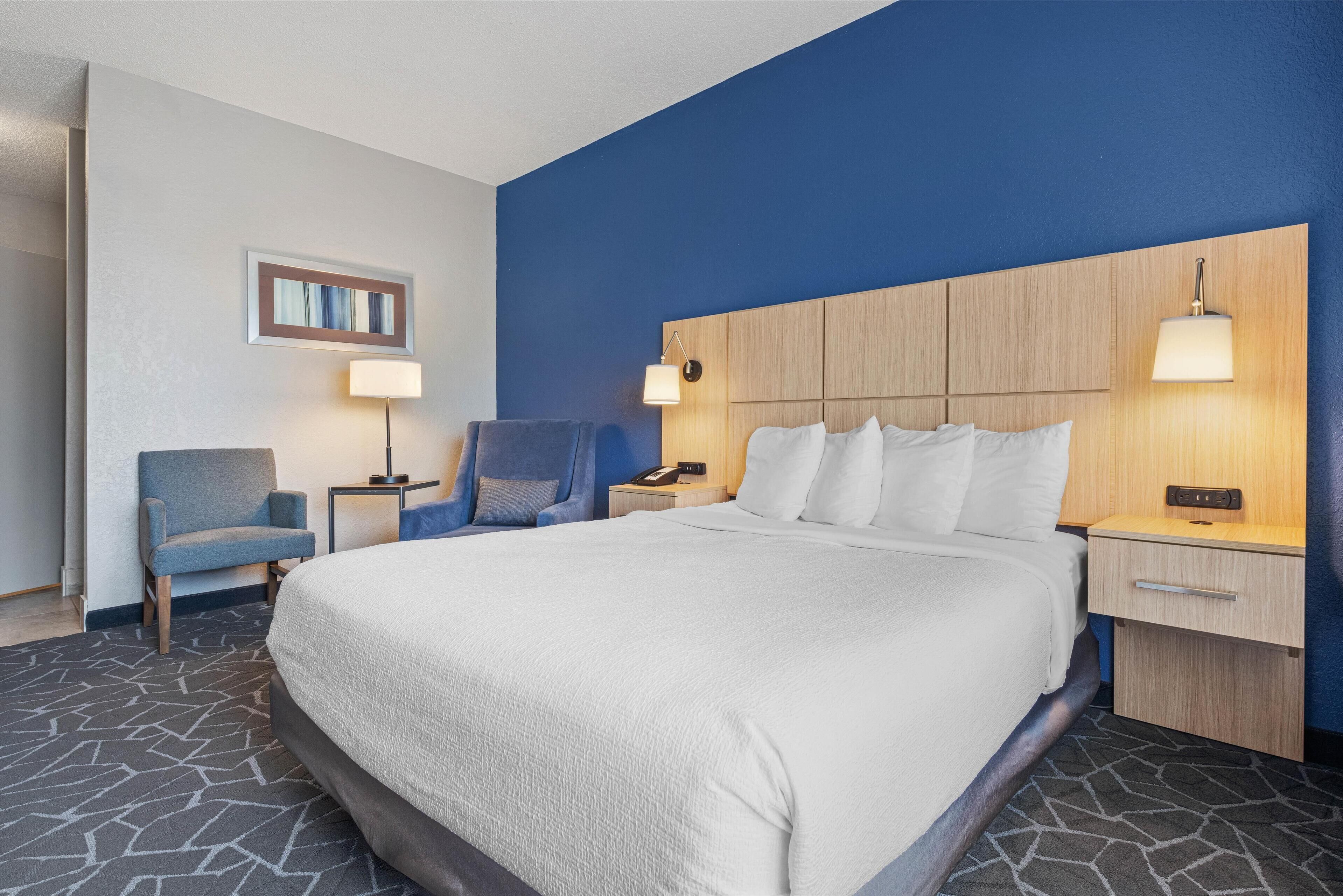 Habitación de hotel con una cama grande, dos sillones, una mesita de noche con lámpara y una luz en la pared.