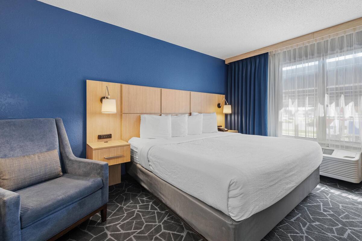 A hotel room with a large bed, a blue armchair, a wooden headboard, two bedside tables with lamps, and a window with curtains.