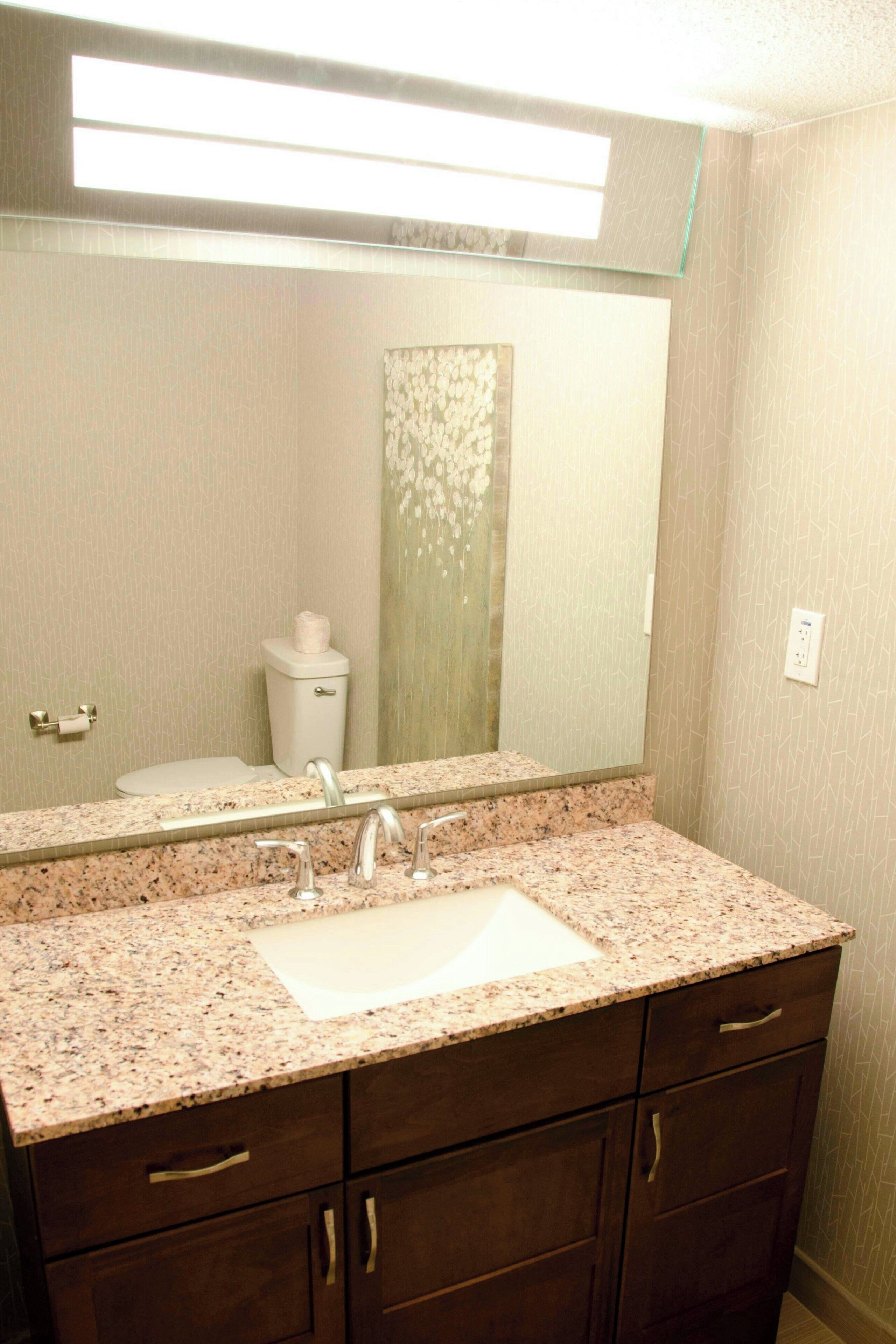 A bathroom with a granite countertop sink, dark wooden cabinets, a toilet, and a mirror above the sink.