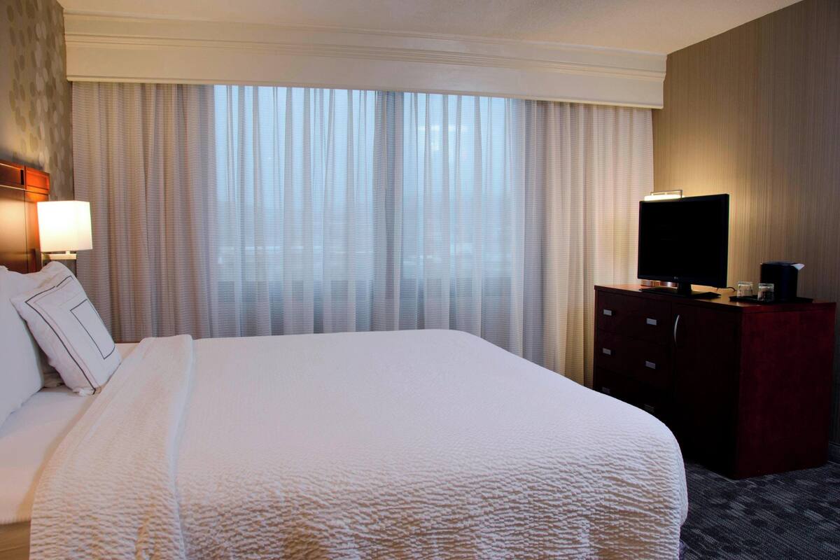 Une chambre d’hôtel avec un grand lit, un téléviseur posé sur une commode et une fenêtre ornée de rideaux légers.