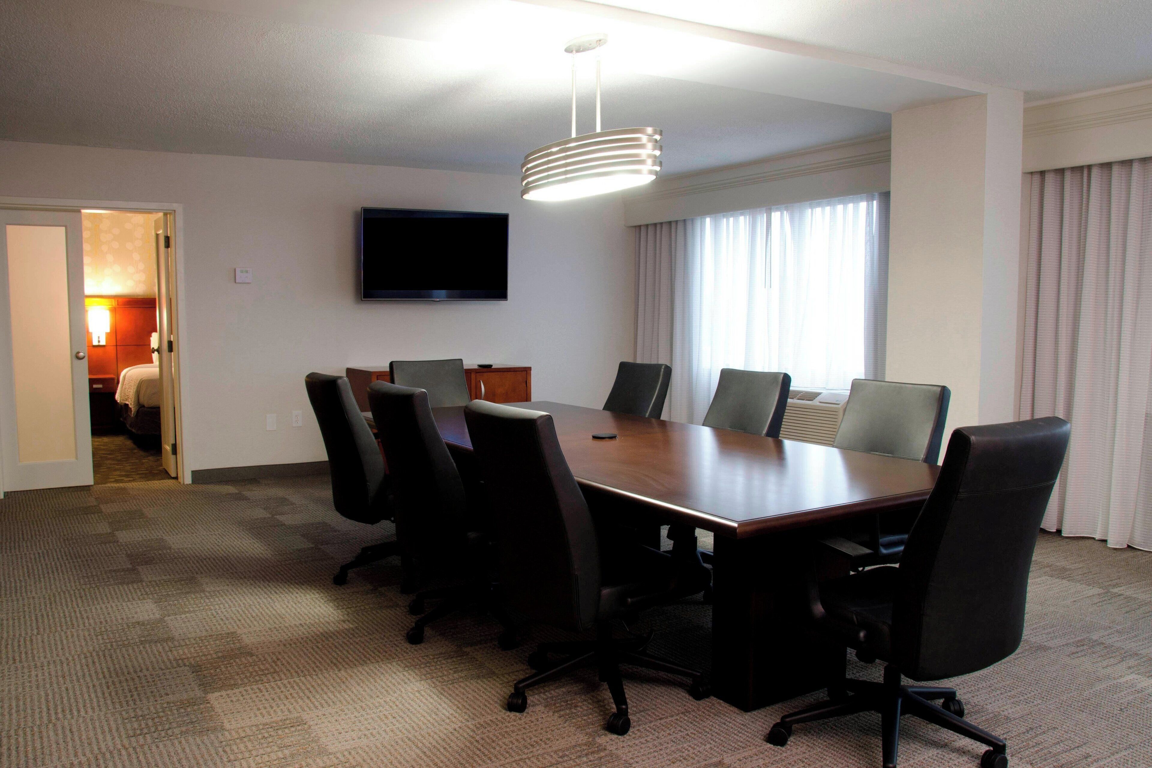 A conference room with a long table, six chairs, a flat-screen TV on the wall, and a large window with curtains.