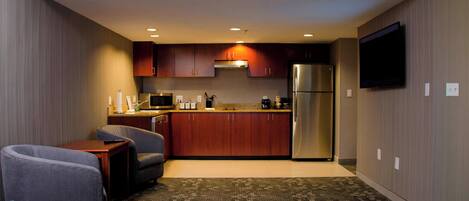 Cocina compacta con alacenas de madera oscura, un refrigerador de acero inoxidable y un televisor de pantalla plana montado en la pared. El área de estar cuenta con dos sillones grises y una mesa pequeña.