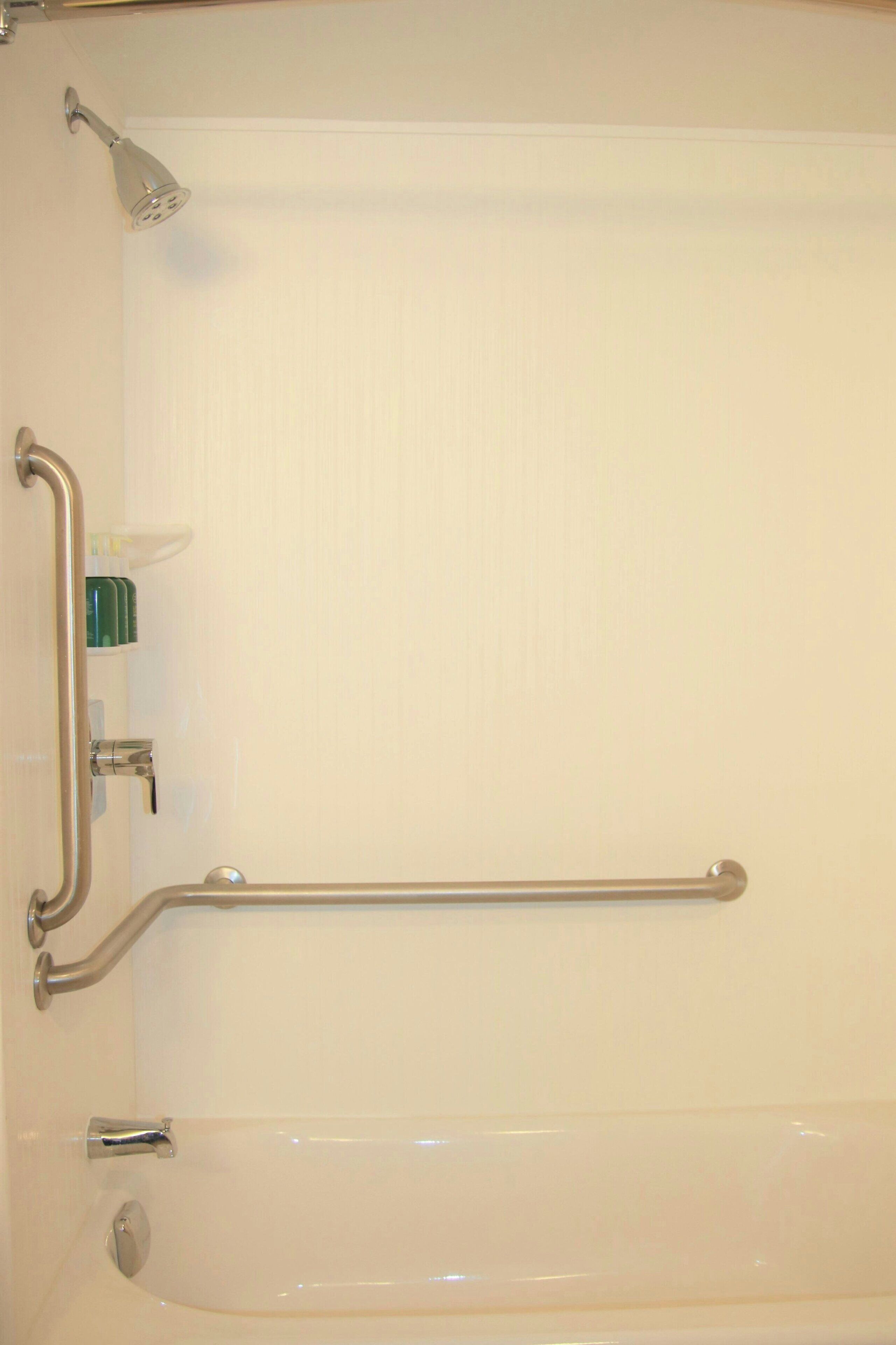 A bathroom with a white bathtub, grab bars, a handheld showerhead, and a soap dispenser.