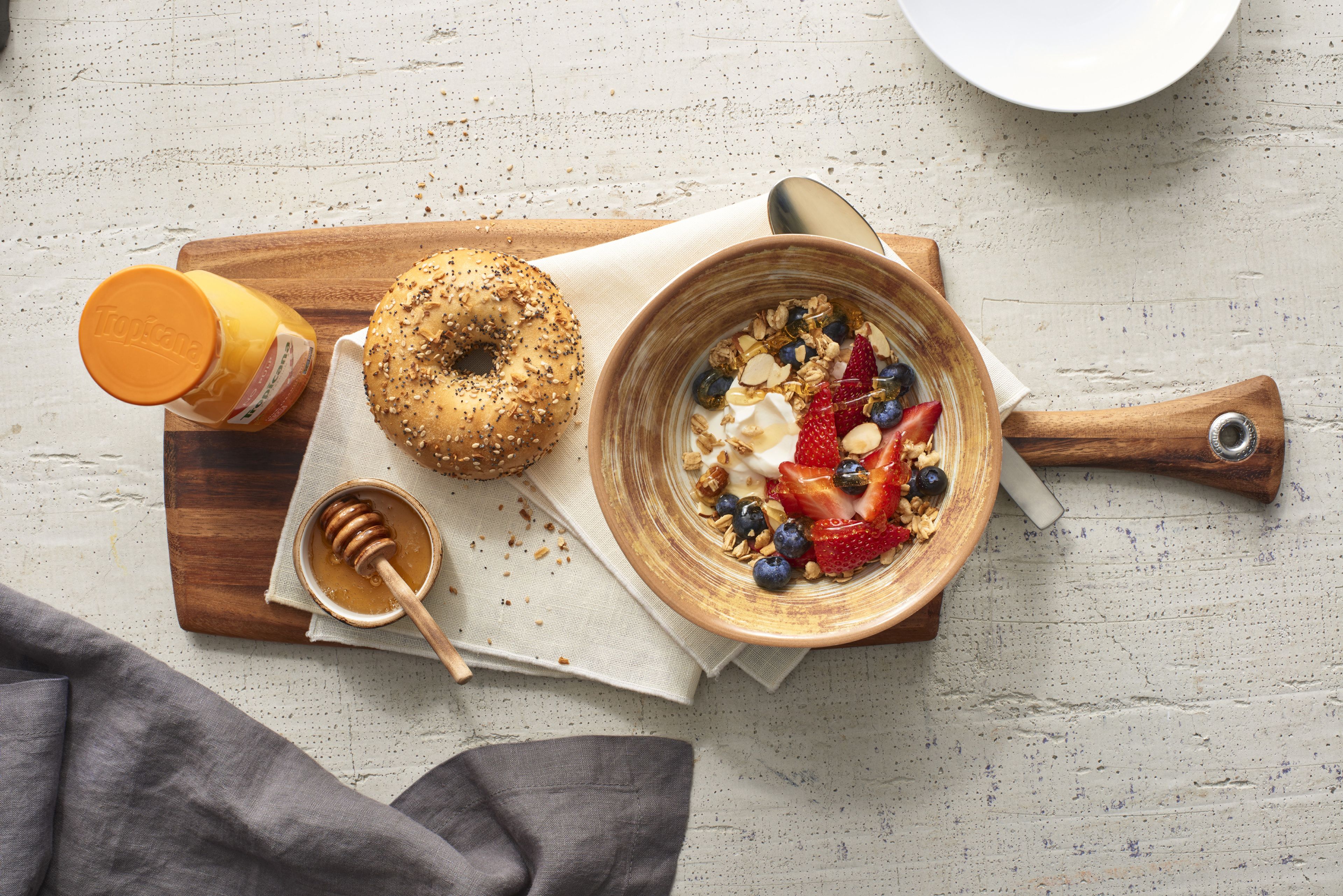 A wooden board with a bowl of yogurt topped with fruits and granola, a honey dipper, and a bagel with seeds.