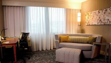 Habitación de hotel con sofá, reposapiés, escritorio, silla y un ventanal con cortinas.