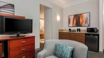 Habitación de hotel con un sillón gris, un televisor de pantalla plana sobre un mueble de madera, un área de cocina con cafetera y un cuadro abstracto grande en la pared.