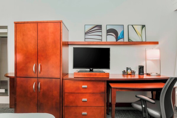 A hotel room with a wooden desk, a flat-screen TV, a lamp, and a cabinet with drawers.