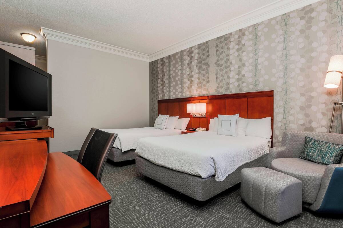 A hotel room with two beds, a desk with a TV, and a seating area with a lamp.