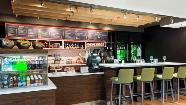 A modern bar area with a wooden counter, stools, a menu board, and a refrigerated display of beverages.