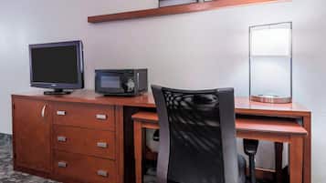 A hotel room with a wooden desk, a television, a microwave, a chair, and a bedside lamp.
