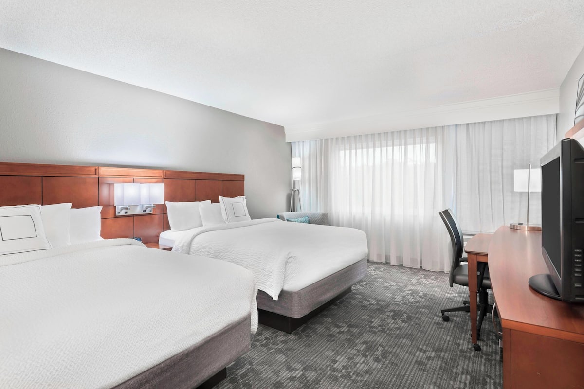 Quarto de hotel com duas camas, uma escrivaninha, uma televisão e uma janela com cortinas.