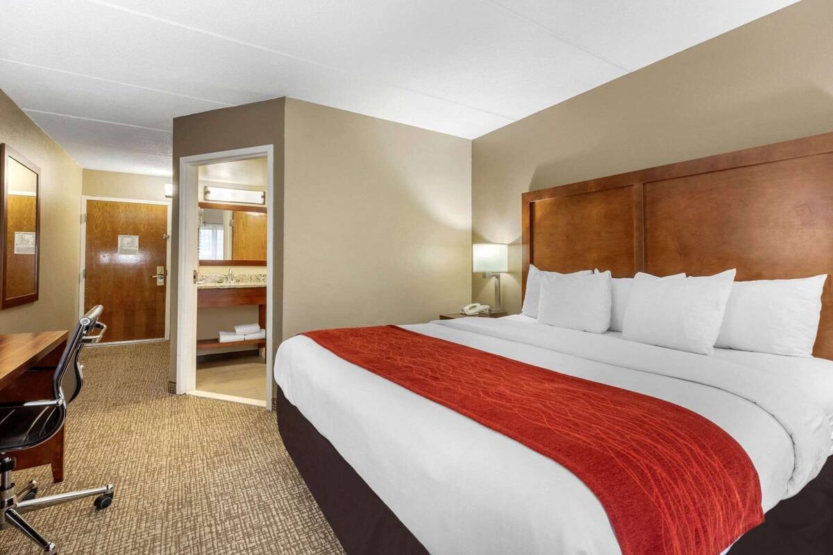 A hotel room with a bed, a desk, a chair, a mirror, and a bathroom visible through an open door.