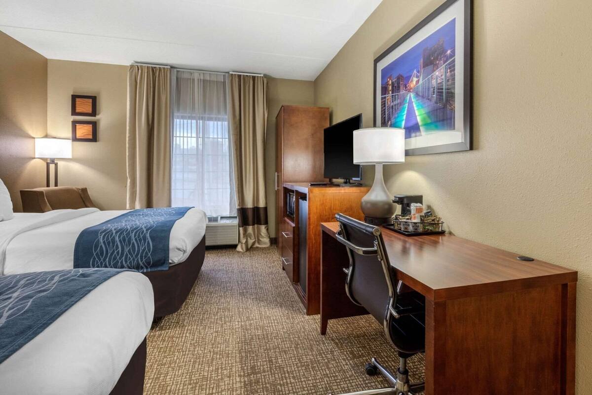 A hotel room with two beds, a desk, a chair, a TV, and a large window with curtains.
