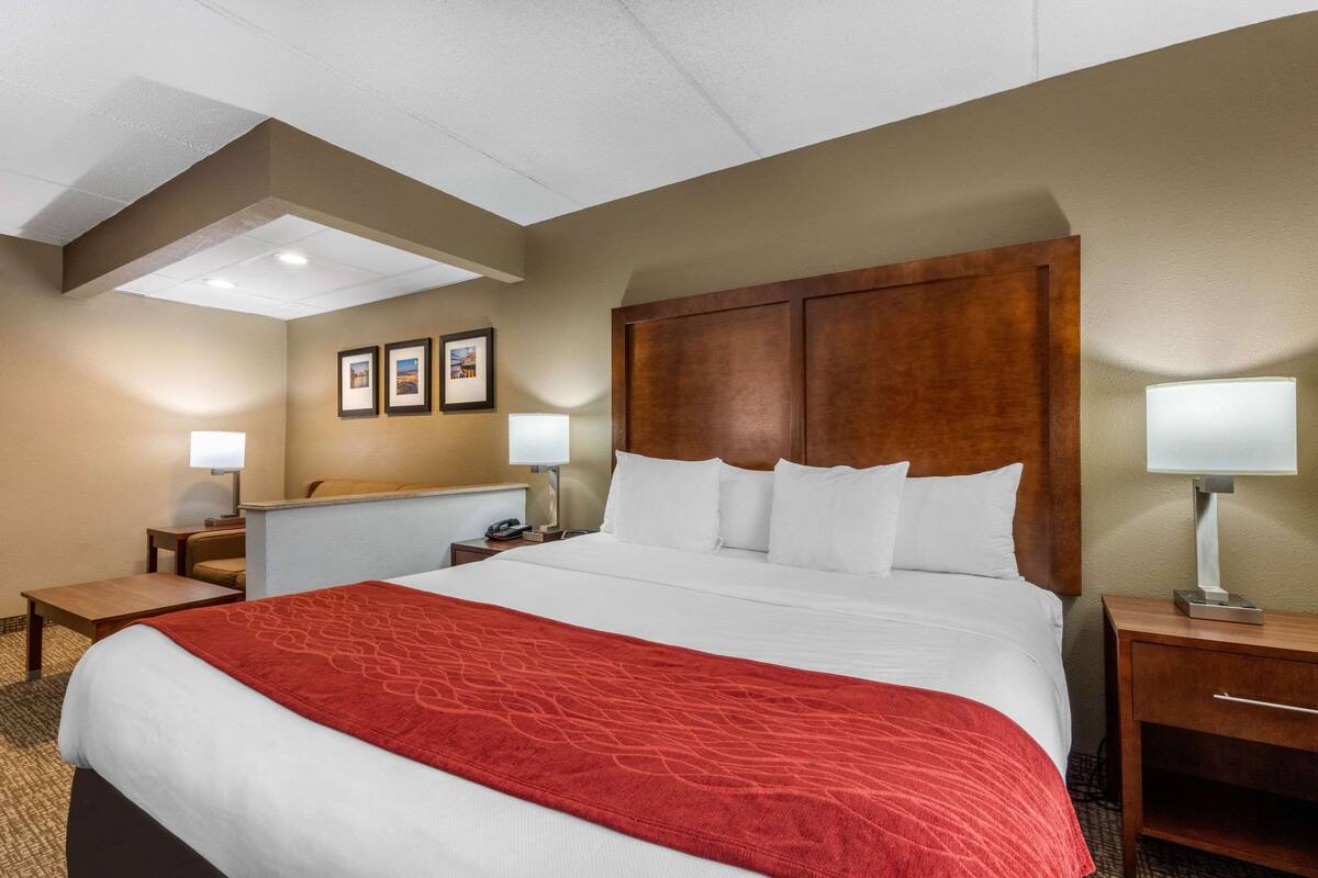 A hotel room with a large bed, a wooden headboard, two bedside tables with lamps, a small desk, and a wall-mounted TV.