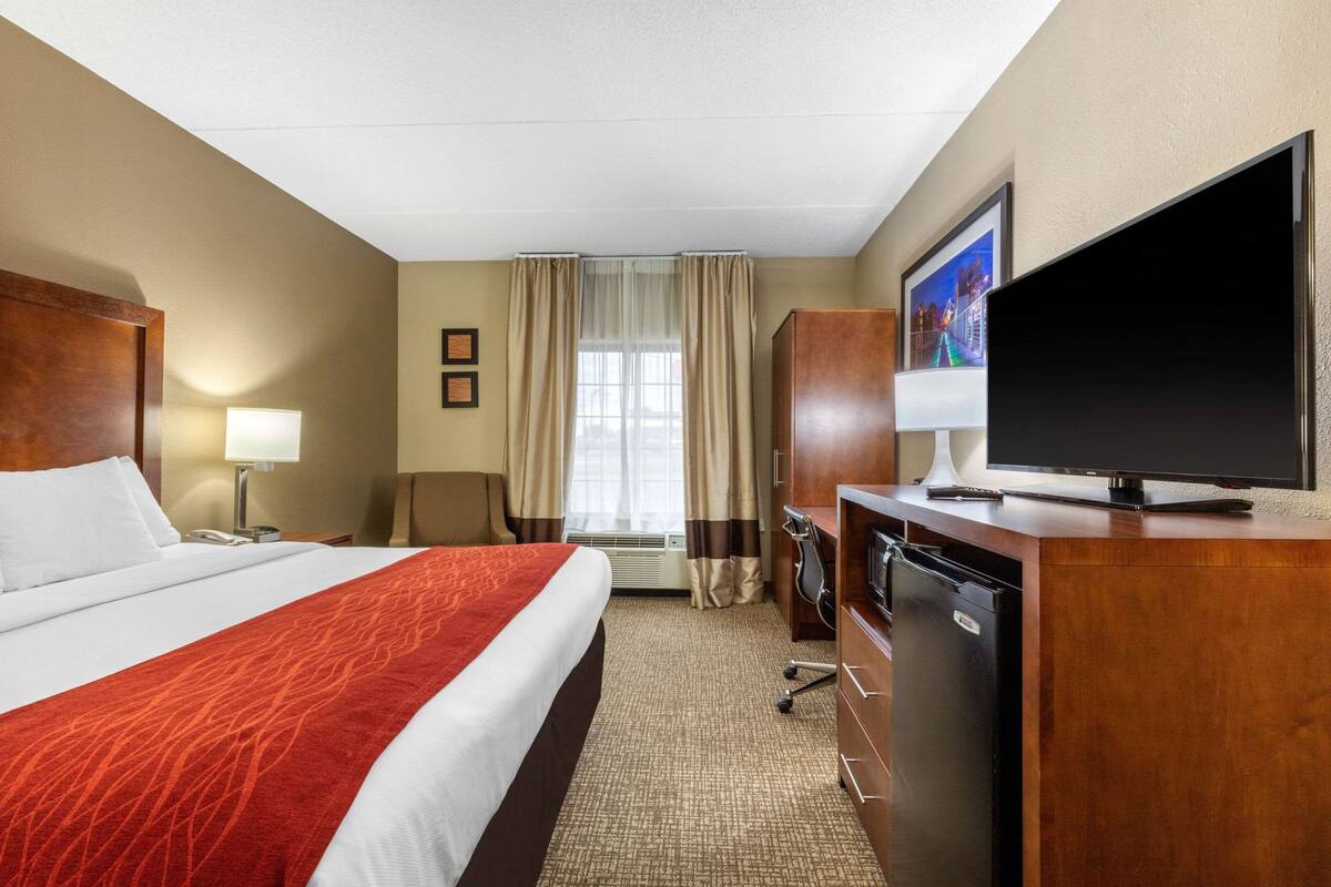 A hotel room with a large bed, a flat-screen TV, a desk with a lamp, a wardrobe, and a window with curtains.