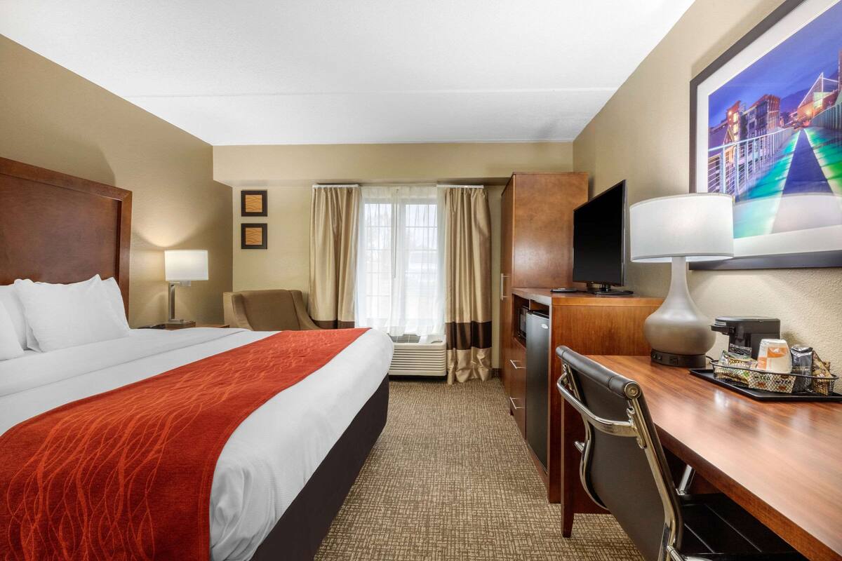 A hotel room with a large bed, a desk with a computer, a chair, a TV, and a window with curtains.