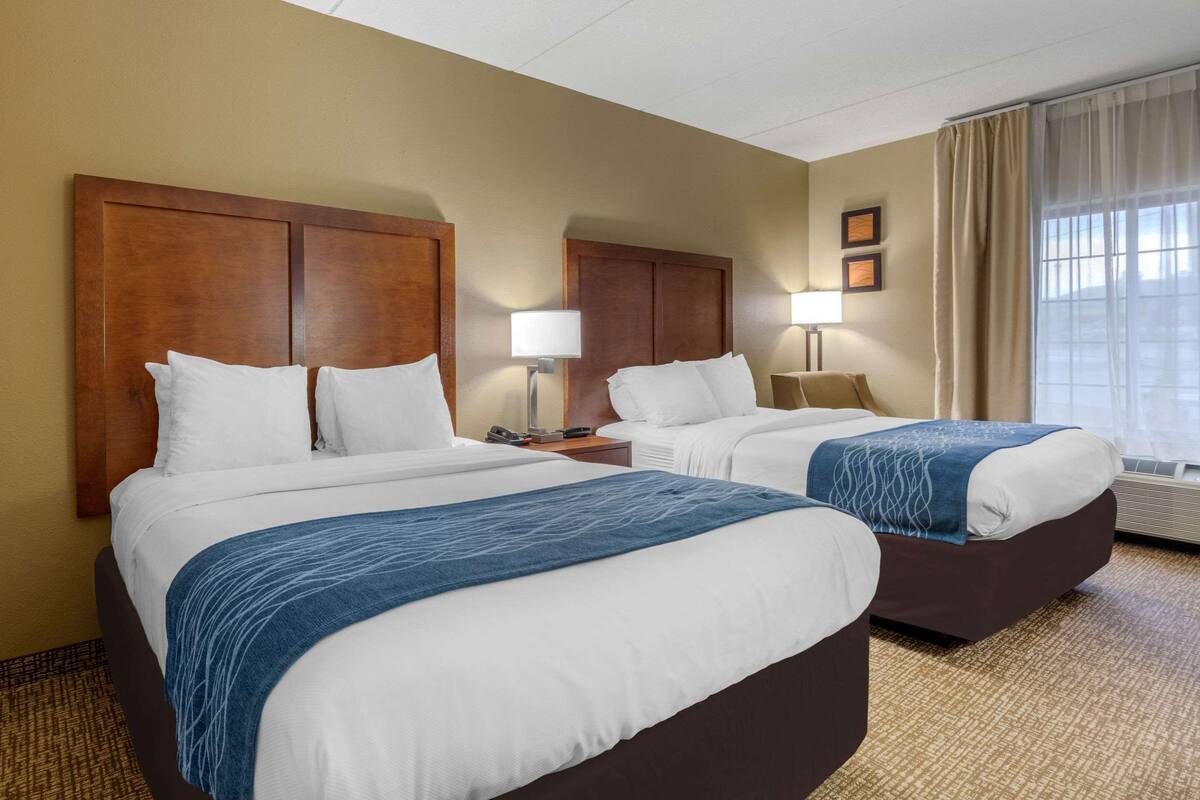A hotel room with two beds, each with a wooden headboard, white bedding, and a blue throw blanket. There are bedside lamps and a window with curtains.