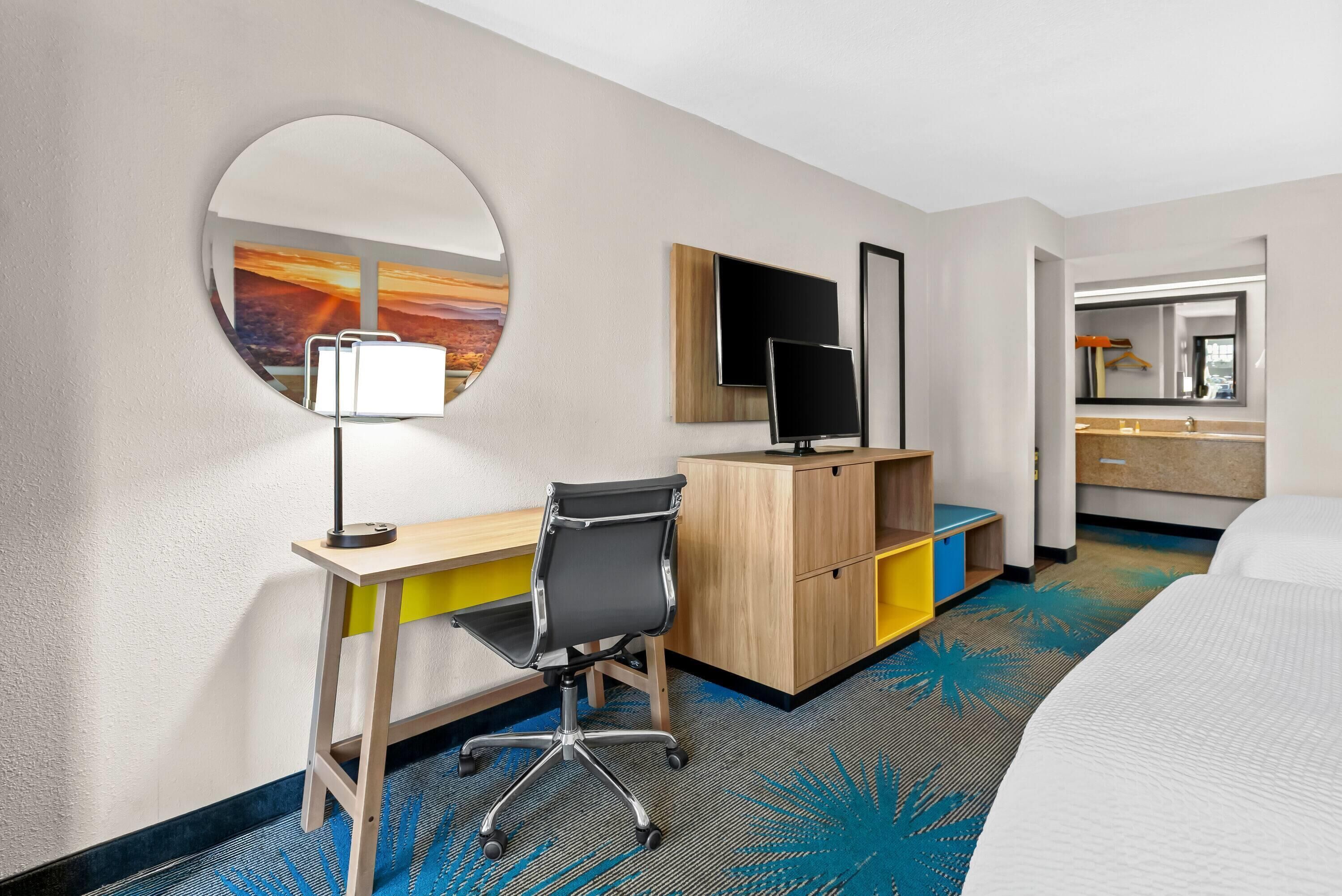 A hotel room with a desk, chair, bed, and a round mirror.