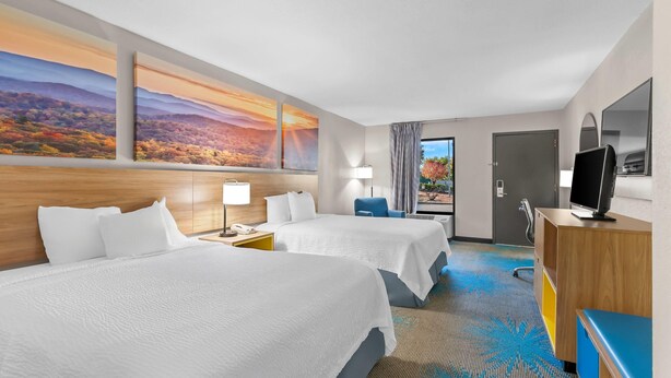Habitación de hotel con dos camas, un escritorio, un televisor y un amplio ventanal con vistas a un paisaje montañoso.