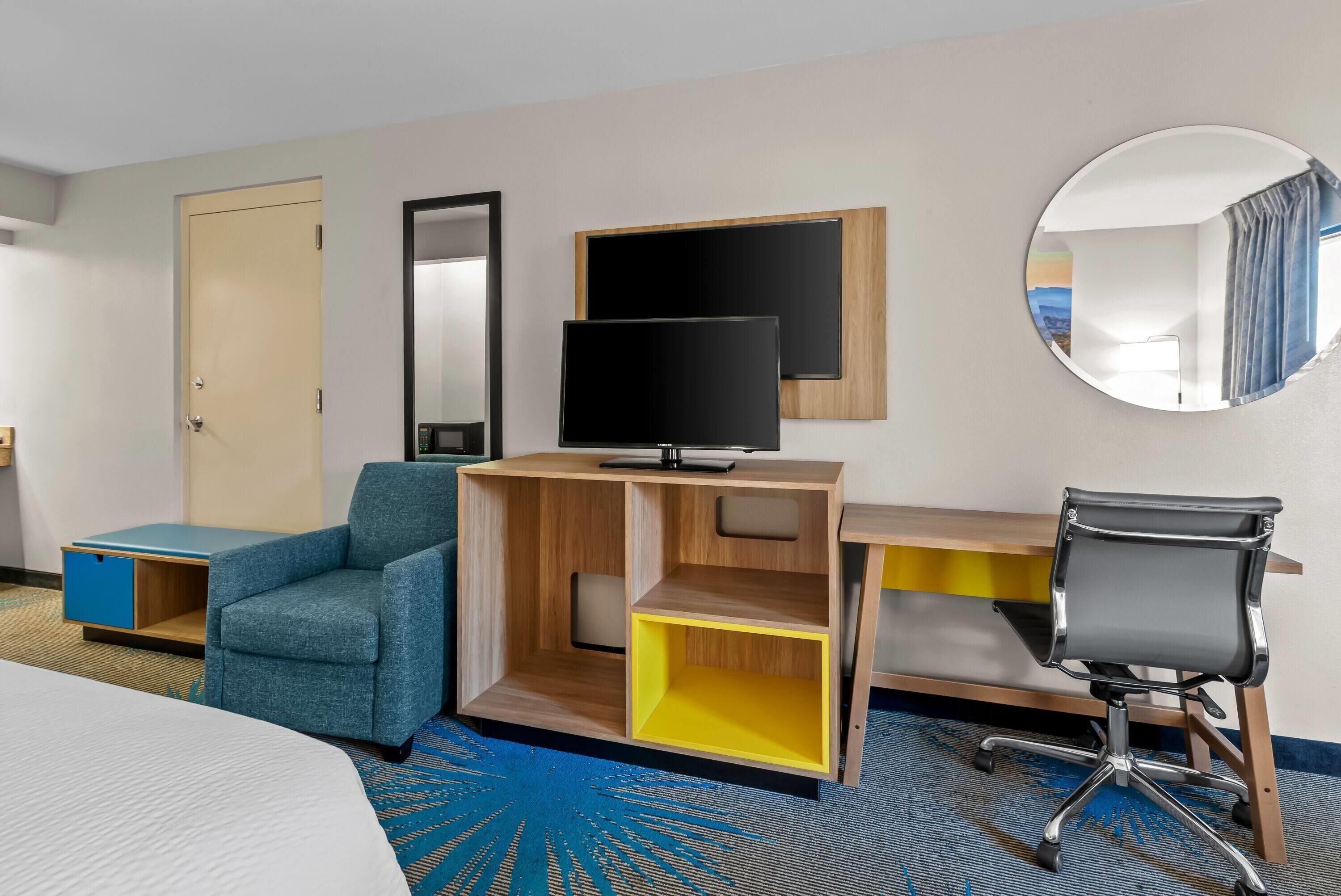 A hotel room with a bed, a desk with a computer, a chair, a TV, a mirror, and a wardrobe.