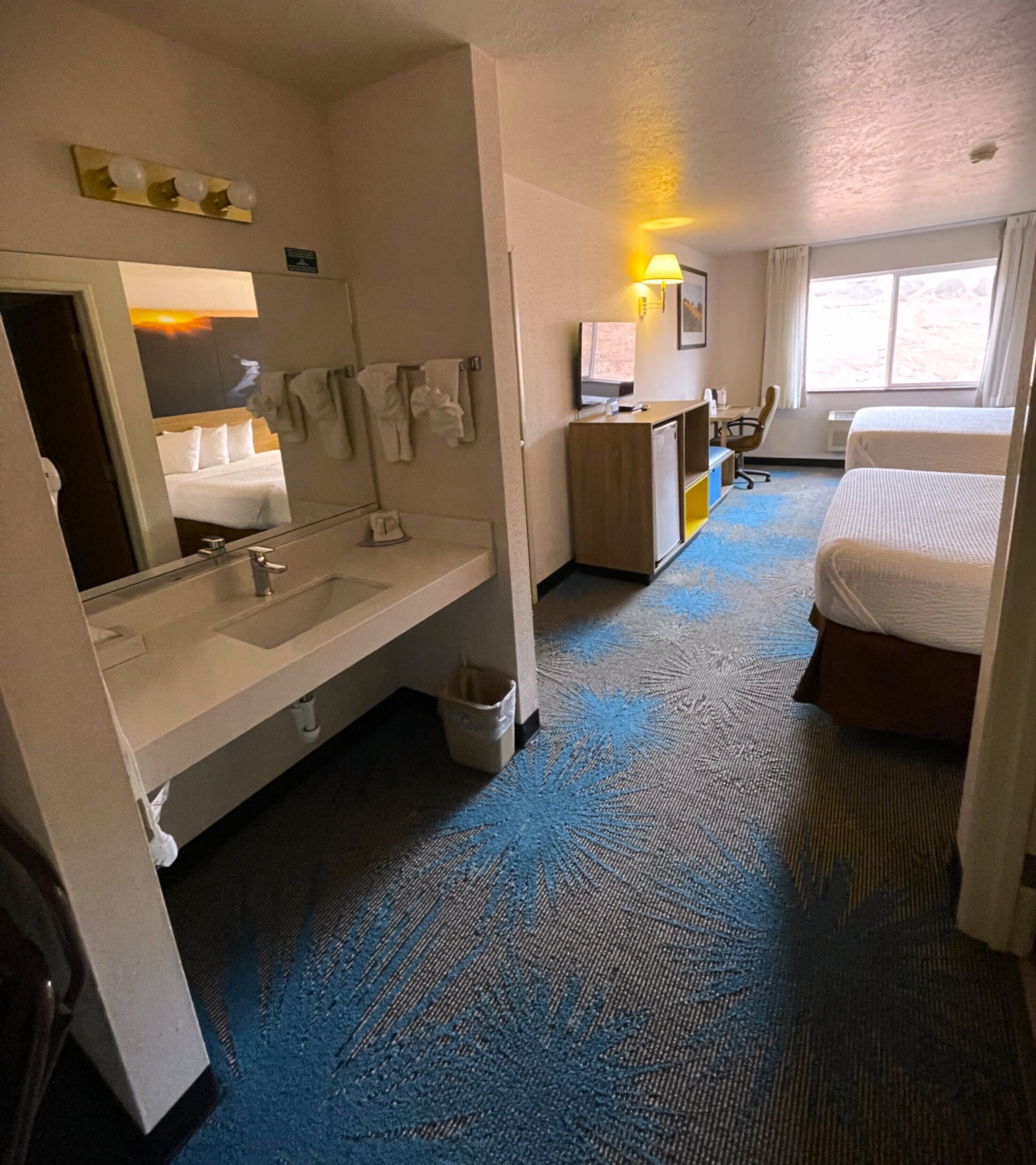 A hotel room with a large mirror, a sink, a desk with a computer, a chair, and a bed.