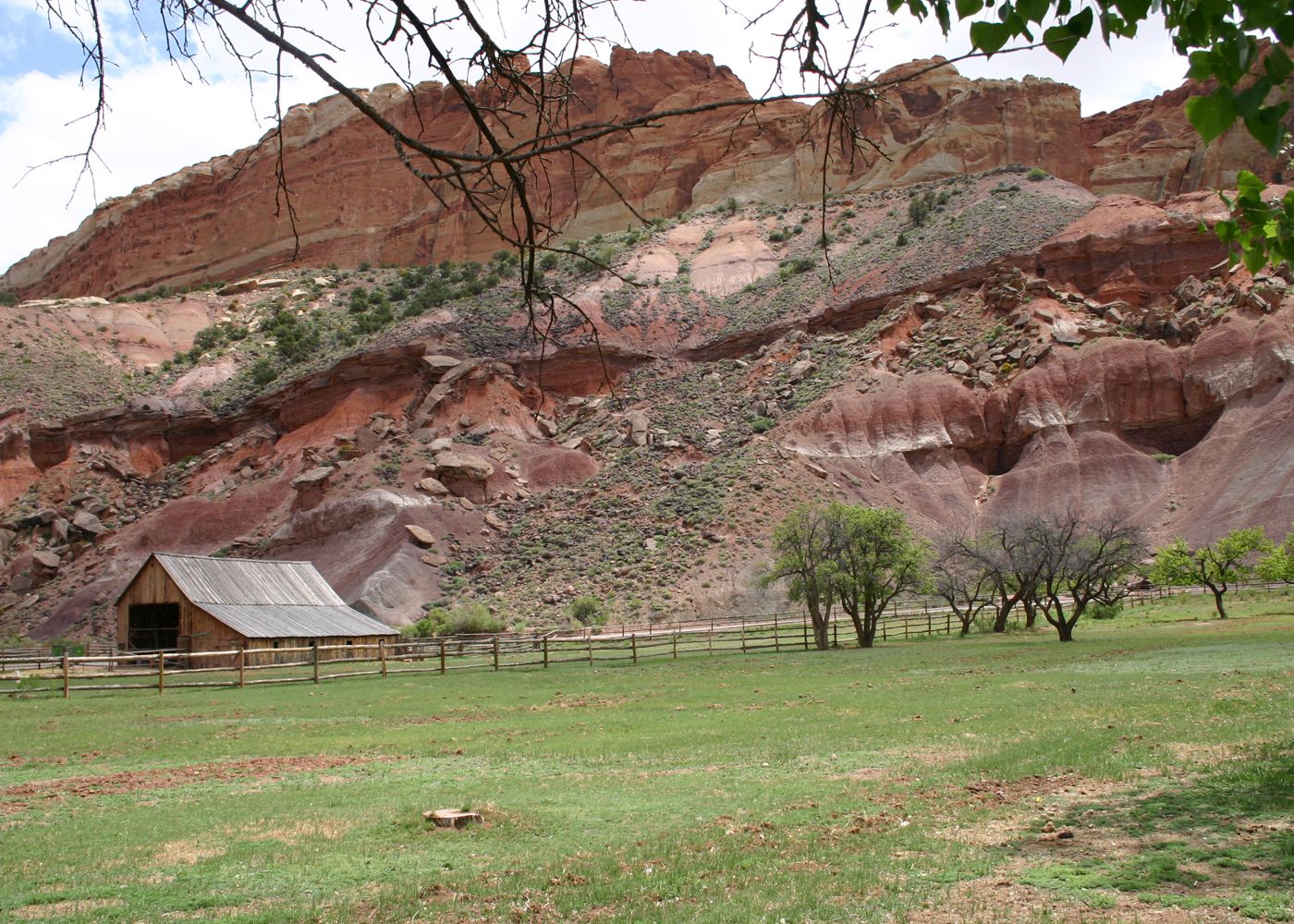 A rustic cabin with a metal roof, surrounded by a wooden fence, set against a backdrop of red rock formations and sparse vegetation.