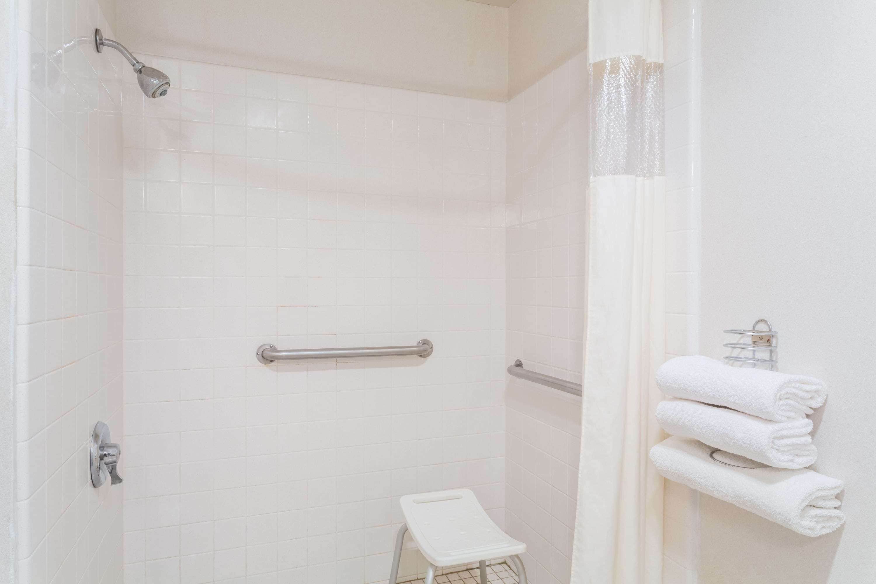 A shower with white tiles, a grab bar, a white step stool, and a towel rack with folded towels.