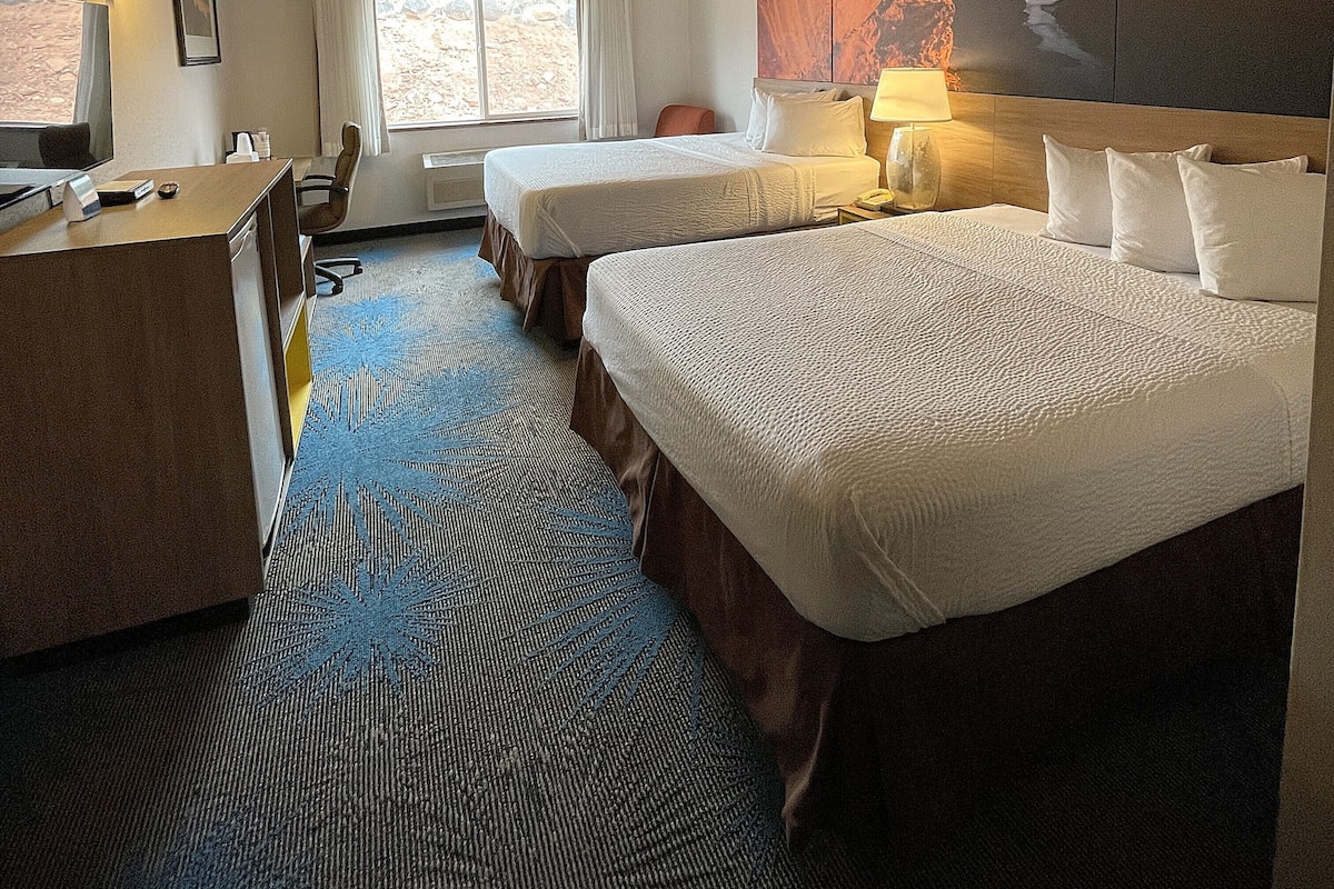 Ein Hotelzimmer mit zwei Betten, einem Schreibtisch, einem Stuhl, einer Lampe und einem großen Gemälde an der Wand.