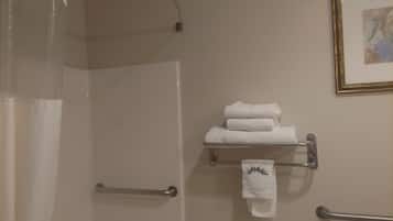 A bathroom with a shower curtain, a towel rack with folded towels, a handrail, and a framed picture on the wall.