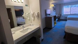 A hotel room with a large mirror, a sink, a bed, a desk with a computer, and a window.