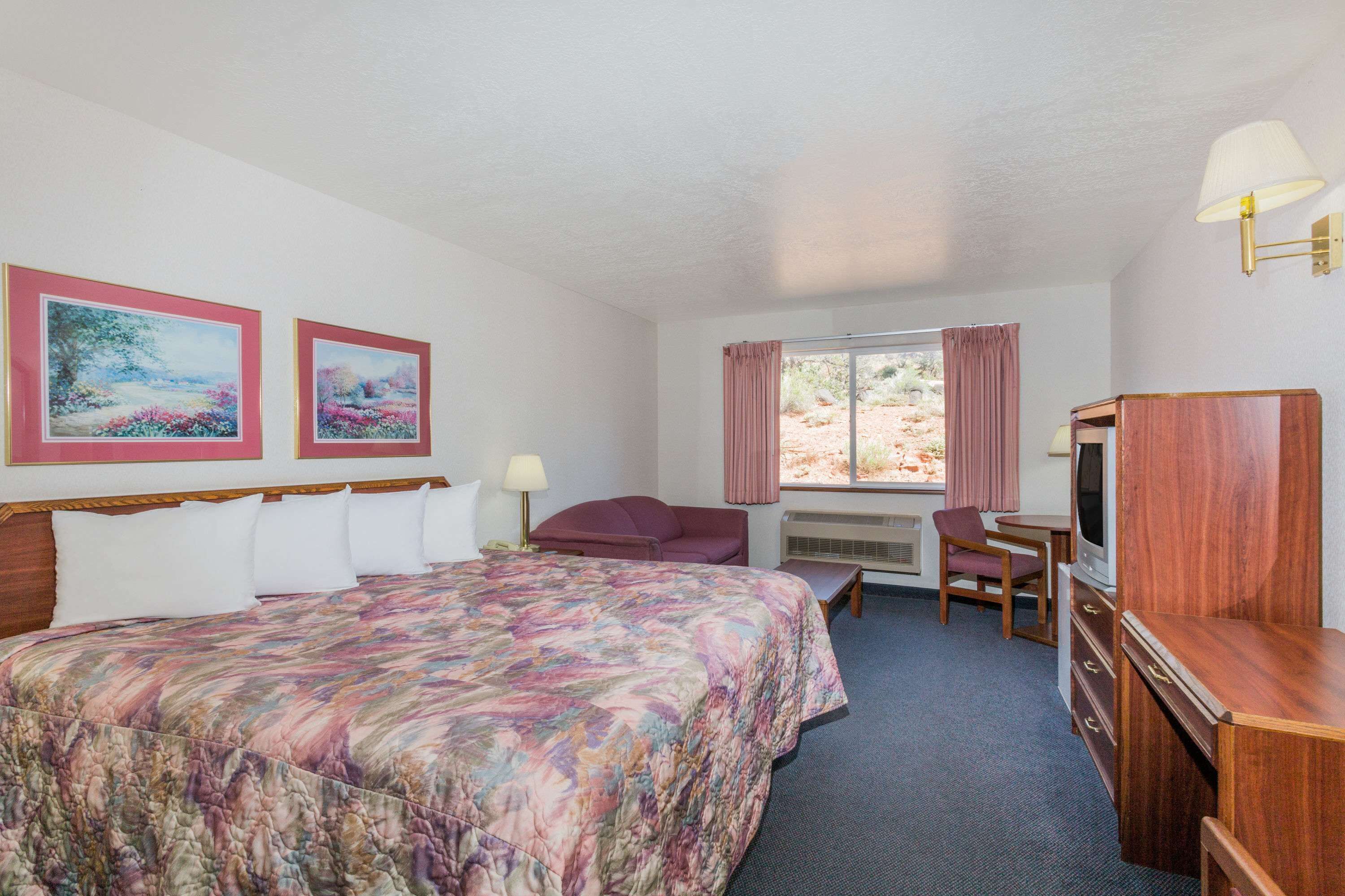 A hotel room with a large bed, a desk with a chair, a television, and two framed paintings on the wall.