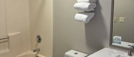 A bathroom with a shower, toilet, sink, and a stack of towels on the wall.