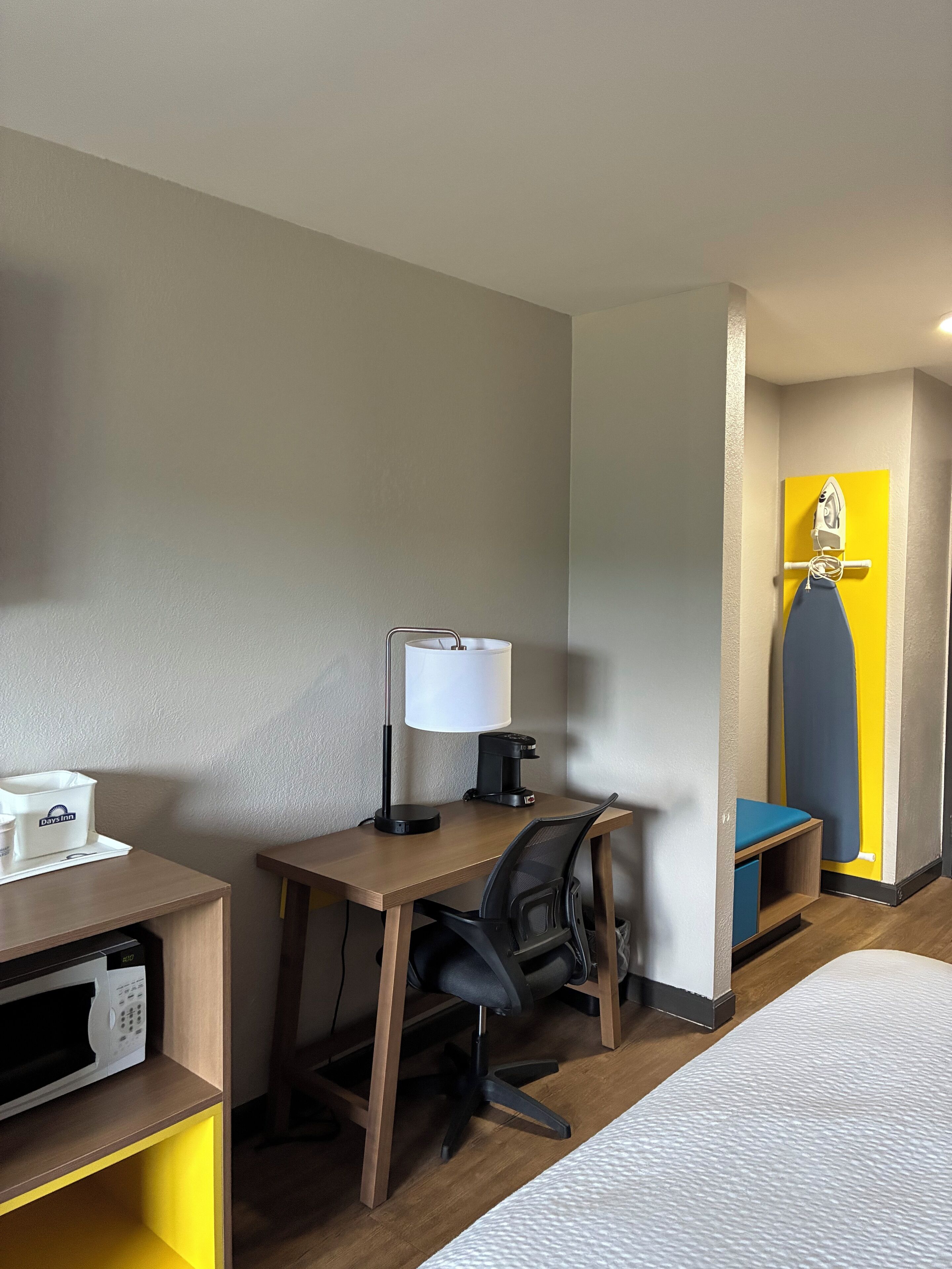 A hotel room with a desk, chair, lamp, coffee maker, and a bed.