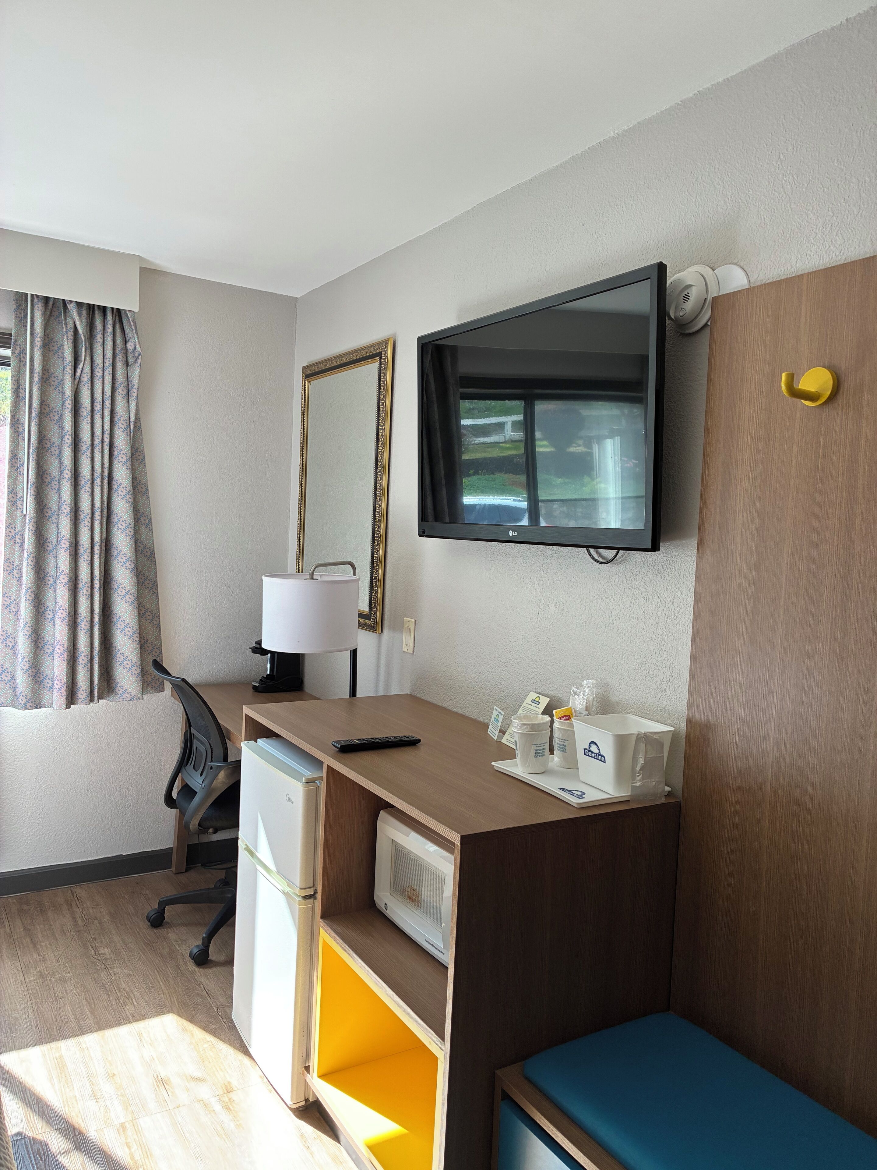 Een hotelkamer met een bureau, een aan de muur bevestigde televisie, een kleine koelkast, een stoel en een bed met een blauw hoofdeinde.